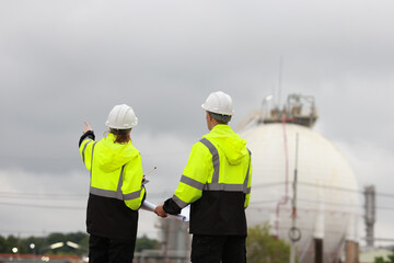 Fototapeta premium Male & Female engineers stand Discussing the building plan at the site, Engineers working outdoors and use walkie-talkies against the Round gas storage tank in the background