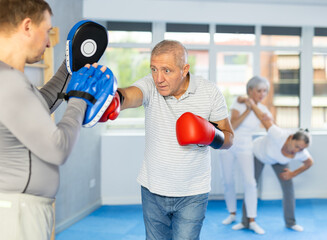 Diligent old man training boxing kicks on punch mitts held by instructor in sports hall