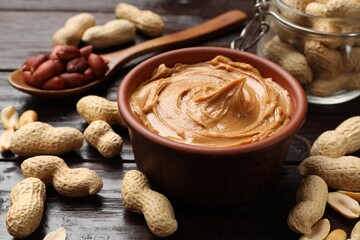 Tasty peanut butter in bowl, groundnuts, jar and spoon on wooden table, closeup