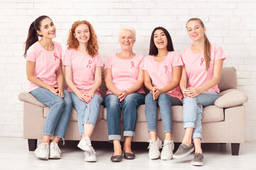 Breast Cancer Survivors. Cheerful Multicultural Women In Pink T-Shirts Smiling To Camera Sitting On Couch Indoors