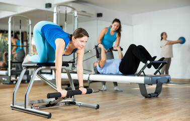Slender middle-aged woman practicing pilates on pedal fitness chair in sports hall during pilates...