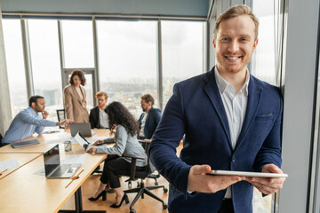 A smiling businessman stands confidently in a modern office setting, holding a tablet while other colleagues work at a nearby table. The office is brightly lit with windows overlooking a city skyline.