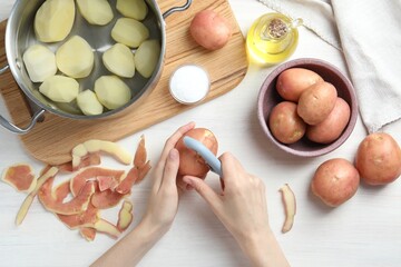 Woman peeling fresh potato with peeler at white wooden table, top view