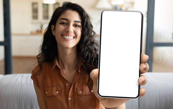 Mobile Advertisement. Happy Beautiful Woman Showing Big Blank Smartphone At Camera While Sitting On Couch At Home, Young Female Demonstrating Phone With White Screen For App Design, Collage, Mockup