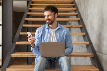 A man sits on wooden stairs in a modern interior. He is holding a smartphone in one hand and has a laptop on his lap. The man is smiling as he looks down at his phone.