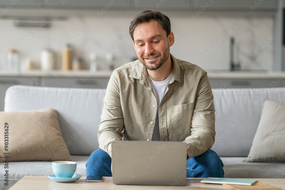 Aufkleber a man sits on a light gray sofa in a modern living room, working on a laptop computer. he is smiling