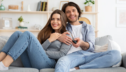 Joyful young couple watching TV together at home, embracing and sitting on couch. Cheerful man and woman switching channels on TV with remote, seeking nice movie for weekend, empty space