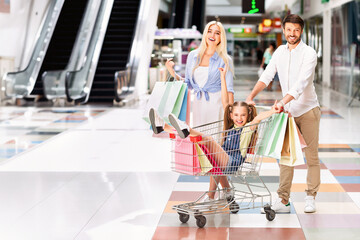 Obraz premium A young girl sits in a shopping cart being pushed by her father, while her mother walks behind with shopping bags. The family is smiling and laughing as they navigate the modern mall