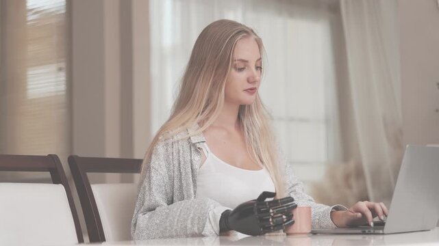 A young woman is using a robotic prosthetic arm hand attached to her body to work on a desk. she is doing computer document and communication work. woman tries to control the smart arm in her home.