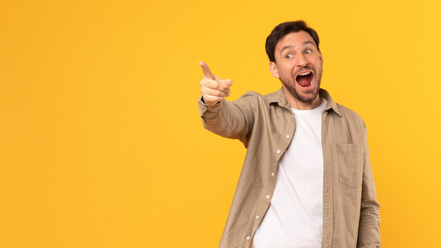 A man in a brown shirt stands in front of a yellow background, pointing his index finger at copy space and looking excitedly in that direction, with his mouth open in a smile.