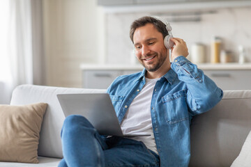 A man sits on a white couch in his living room, wearing a denim shirt, jeans, and headphones. He...