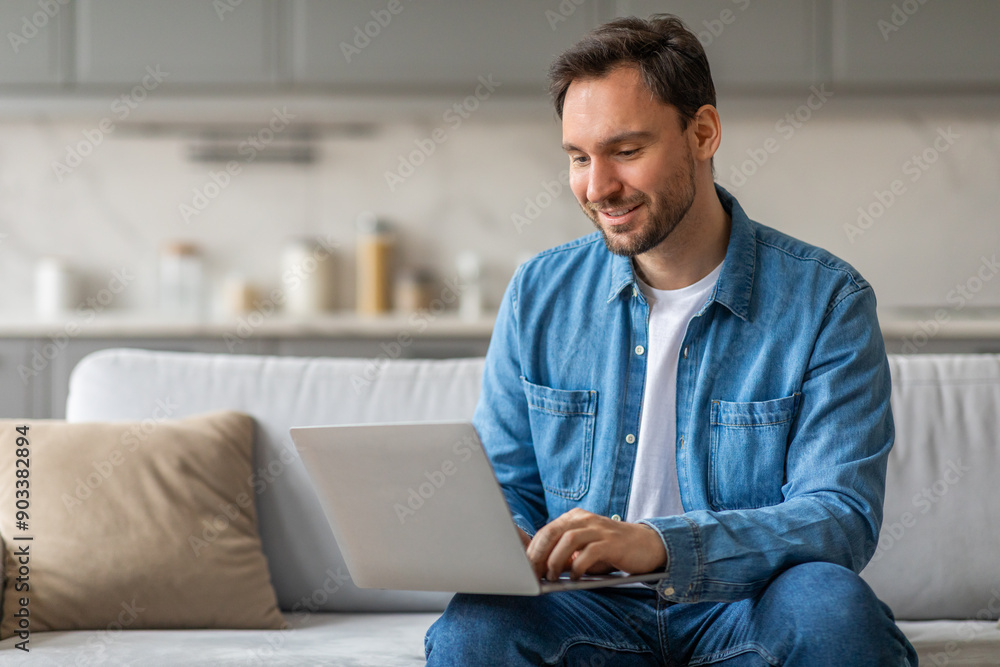 Aufkleber man in a blue button-down shirt sits on a white couch in a living room. he is smiling and working on