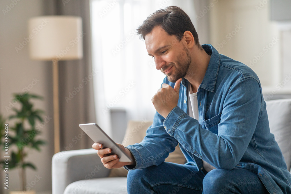 Poster a man wearing a blue denim shirt sits on a gray couch in a living room. he is using a tablet compute
