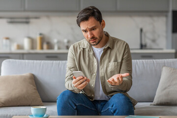A man sits on a couch in his living room, holding his phone with a confused expression on his face....