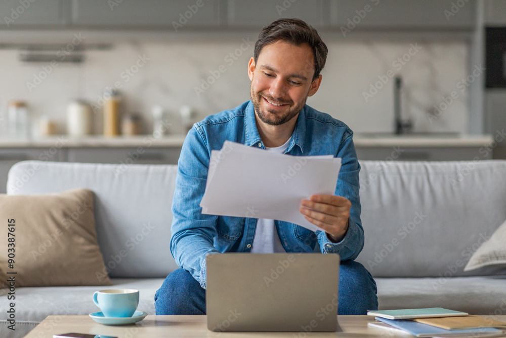 Poster A man is sitting on a sofa, smiling as he reviews some documents while using a laptop computer. He is wearing a blue denim shirt, and the sofa is in a home living room.