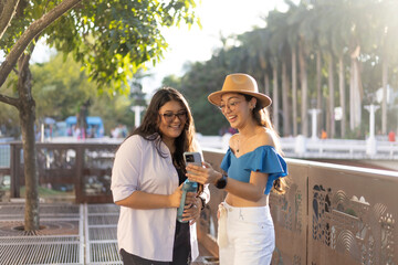 Two young girls with long hair and glasses in an urban environment looking at their mobile phones, enjoying themselves and smiling together.