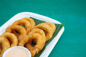Tasty onion rings served on biodegradable tray with green leaf and sauce.