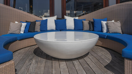A place to relax. A semicircular wicker sofa with a soft mattress on the seat. A lot of pillows are stacked in a row. A round white table in the center.
