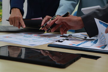 A businessman sits at a desk in an office, discussing documents with coworkers. Using a laptop, he engages in teamwork and communication, focusing on finance and corporate success