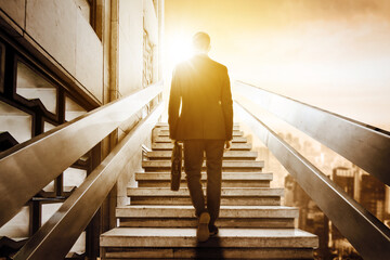 Rear view of male manager carrying a suitcase while walking upward on the stairs with modern city background