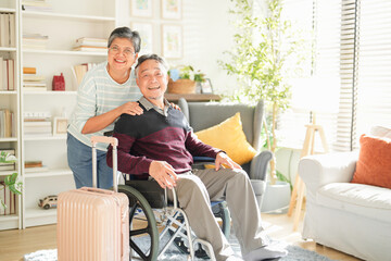 An elderly couple is excited and ready for a travel adventure, with the man in a wheelchair and a suitcase beside them. They share a joyful moment in their cozy living room, showcasing their love. © ztony1971