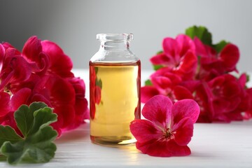 Bottle of geranium essential oil and beautiful flowers on white wooden table, closeup