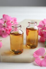 Bottles of geranium essential oil and beautiful flowers on white table, closeup