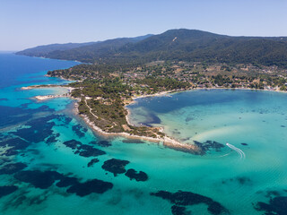 Aerial view of an island on Sithonia Peninsula, Greece, with beaches and rocks, surrounded by crystal-clear waters. Yachts and boats anchor in turquoise waters. Nearby islets and reefs visible