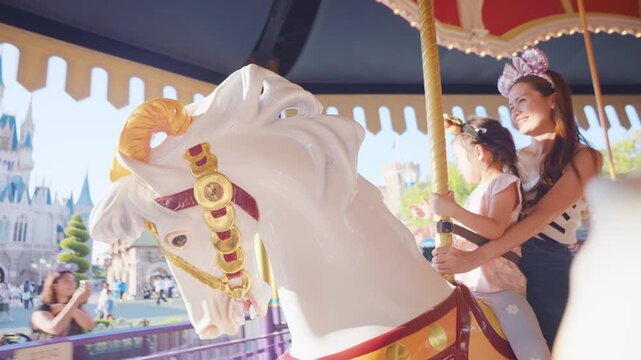 Asian mother and adorable kid daughter riding carousel at theme park. 