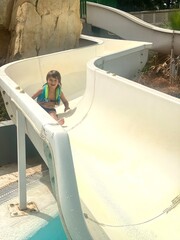 A smiling 3-year-old Caucasian boy enjoying a water slide on a sunny day at the pool.
