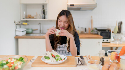 Young asian woman enjoying to having breakfast with eating delicious sandwich while cooking fresh vegetables salad and homemade snack for health food in modern kitchen with healthy lifestyle in home