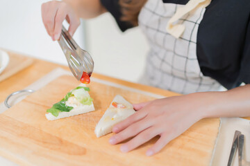 Young asian woman using tongs to taking tomatoes on the bread and making delicious sandwich for healthy breakfast food to preparing homemade snack in modern kitchen with healthy lifestyle at home