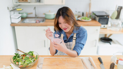 Young asian woman enjoying to eating delicious yogurt after preparing fresh vegetables salad and...