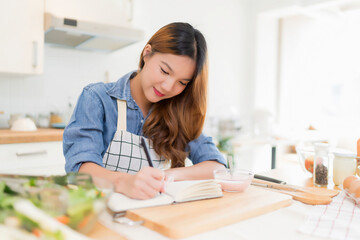 Young asian woman writing food recipes in notebook and eating yogurt while preparing fresh...