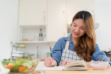 Young asian woman writing food recipes in notebook and eating yogurt while preparing fresh vegetables salad and cooking healthy breakfast food in modern kitchen with healthy lifestyle at home