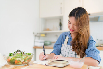 Young asian woman taking notes about food recipes in notebook and eating yogurt while preparing vegetables salad and cooking healthy breakfast food in modern kitchen with healthy lifestyle at home