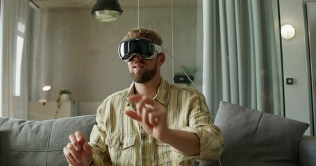 A man in a virtual reality headset moves his hands while sitting on a sofa in a home interior, daylight - Powered by Adobe