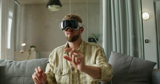 A man in a virtual reality headset moves his hands while sitting on a sofa in a home interior, daylight