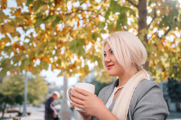 A blonde woman is sitting on a bench and drinking coffee. She is wearing a gray jacket and scarf.