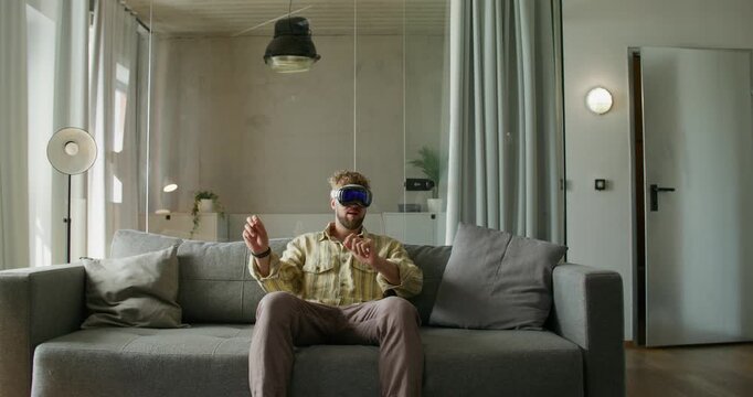 A man in a virtual reality headset moves his hands while sitting on a sofa in a home interior, daylight