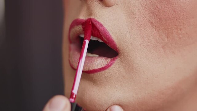 Young performer in drag queen makeup using a brush to apply the lipstick in the dressing room during preparation for the performance close up