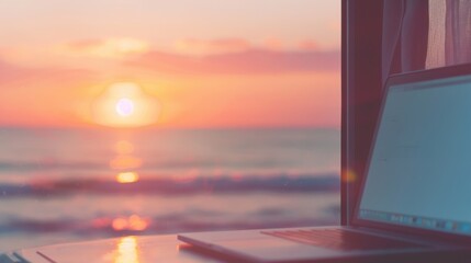 A laptop sits on a desk beside an open window, with a beautiful sunset casting colors over the ocean