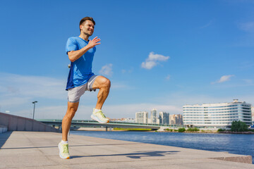 A man in a blue athletic shirt performs a high knee exercise by a riverfront, with a city skyline...