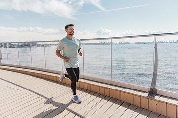 A man in athletic wear jogs along a waterfront boardwalk, smiling and enjoying the sunny day.