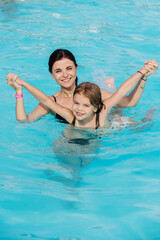 A young woman and her daughter swim in a transparent pool, smile and enjoy the time spent together. The scene captured summer holidays, family happiness, joy and carefree fun, perfect for the holidays