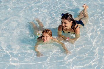 A young woman and her daughter swim in a transparent pool, smile and enjoy the time spent together. The scene captured summer holidays, family happiness, joy and carefree fun, perfect for the holidays