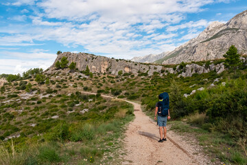 Obraz premium Hiker man with baby carrier backpack on idyllic hiking path to Tucepi town along Makarska Riviera, Dalmatia, Croatia, Europe. Majestic Biokovo mountains rise up against dramatic sky. Dinaric Alps