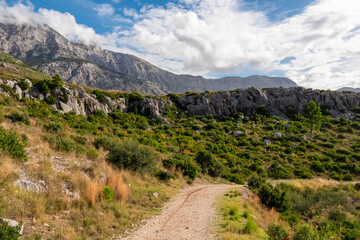 Winding idyllic hiking path to Tucepi town along Makarska Riviera, Split-Dalmatia, Croatia, Europe. Majestic Biokovo mountain range rising up against dramatic sky. Scenic landscape of Dinaric Alps