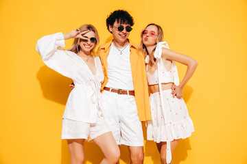 Fototapeta premium Group of young three stylish friends posing in studio. Fashion man and two cute female dressed in casual summer clothes. Smiling models having fun. Cheerful women and guy near yellow wall, sunglasses