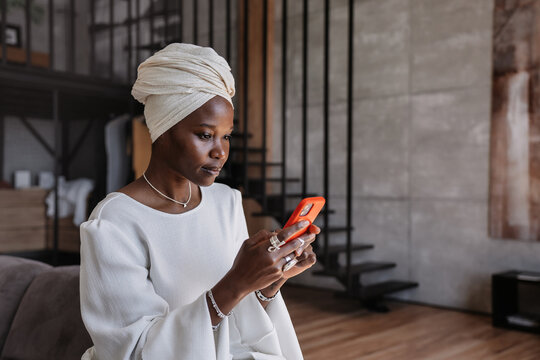 focused woman in a white outfit and head wrap sitting in a modern living space, intently using her smartphone, blending traditional and contemporary elements
